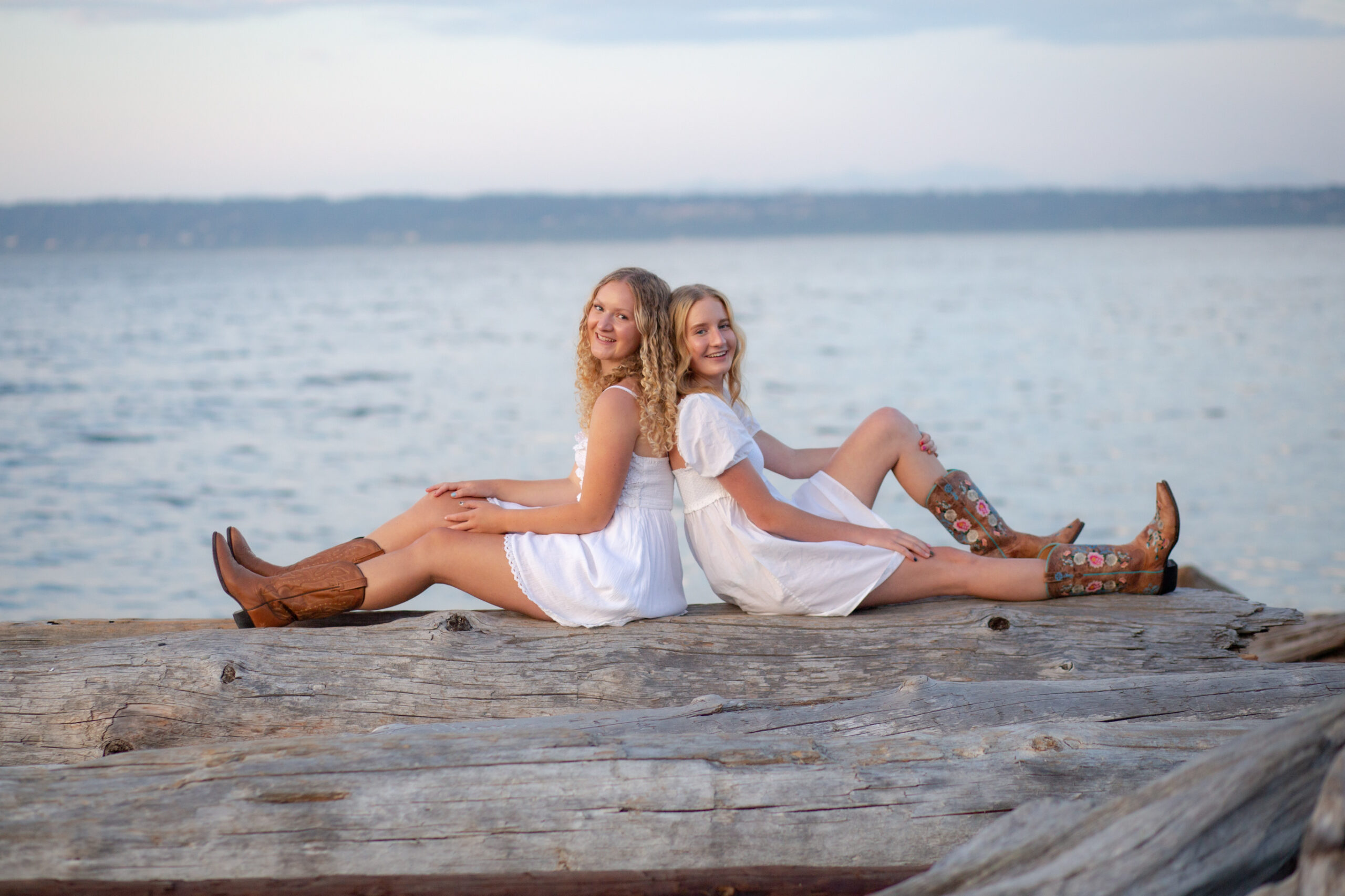 Two high school girls in white dresses and brown cowboy boots. They’re sitting back to back on driftwood in front of the water on Bainbridge Island.