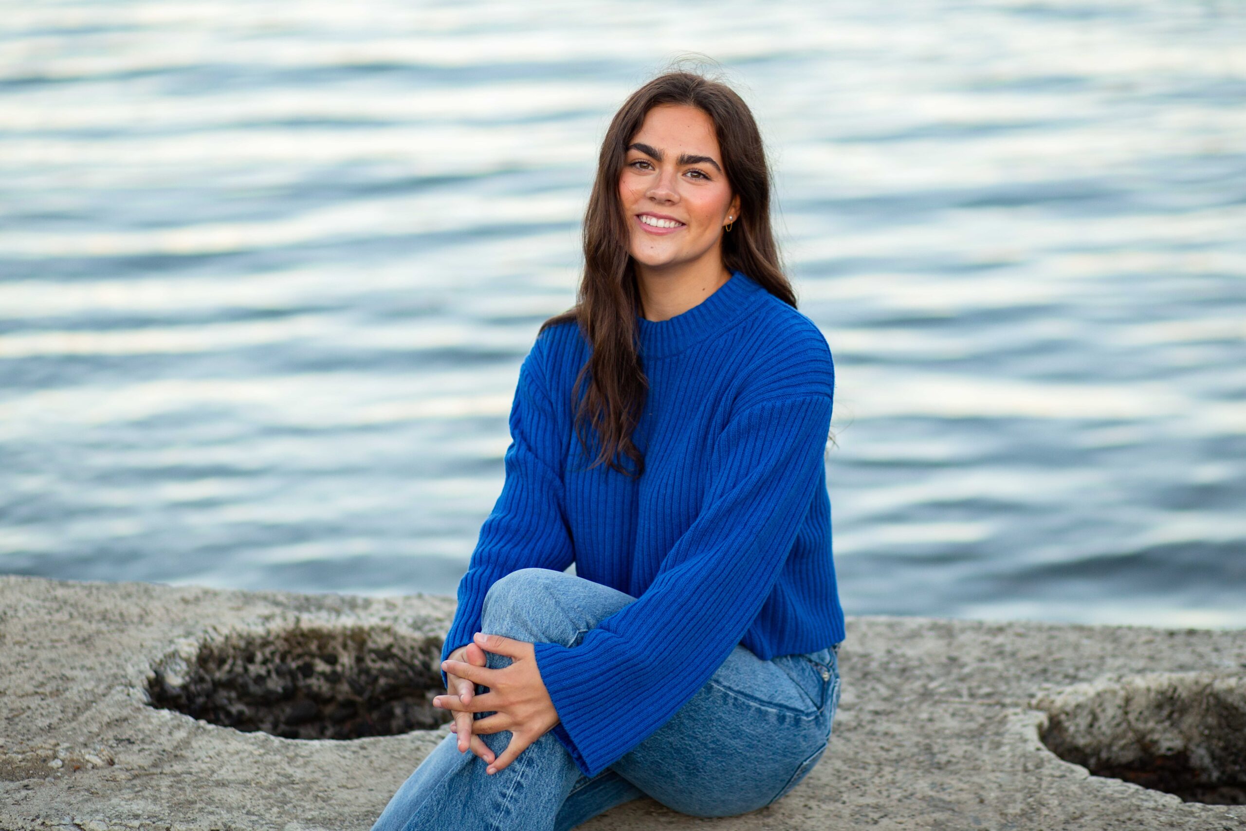 High school senior with long, dark hair wearing a blue sweater and jeans in front of the water on a cement platform.
