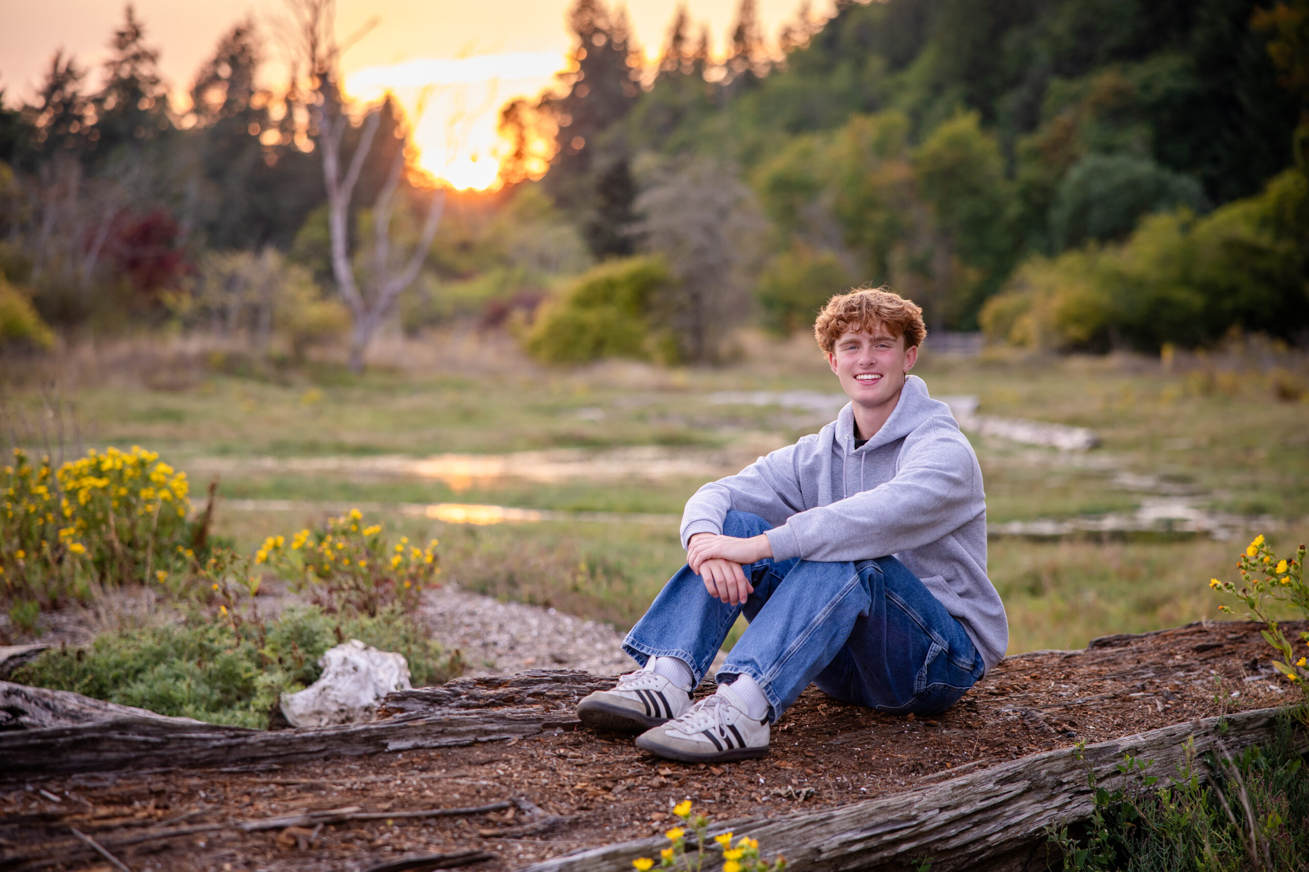 High schooler sitting on wood in front of an open field. He’s wearing a gray hooded sweatshirt and blue jeans.